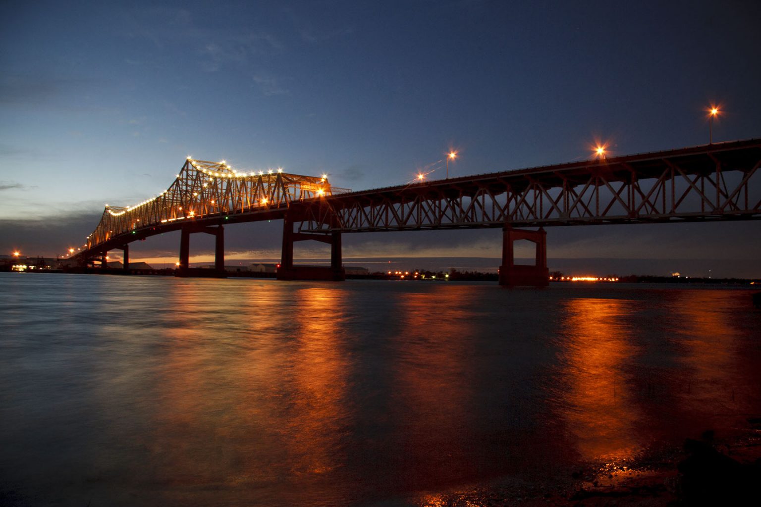 Illuminated steel truss bridge over calm water at dusk with orange reflections.